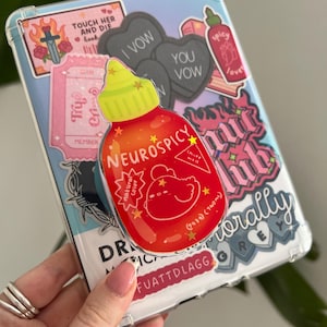May include: A clear phone case adorned with various stickers. A prominent red cartoon hot sauce sticker reads "NEUROSPICY". Other stickers feature hearts, text, and a pink club sticker. The case is held against a blurred green background.