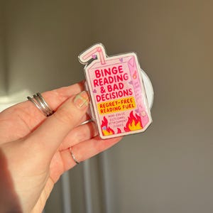 May include: A pink juice box-shaped sticker with the text "Binge Reading & Bad Decisions" and "Regret-Free Reading Fuel." The sticker features flames and hearts, suggesting a fun, book-themed design.
