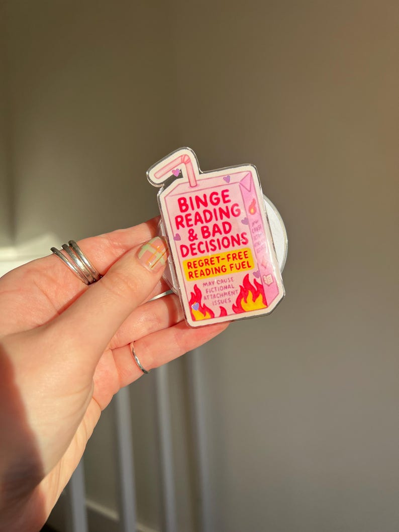 May include: A pink juice box-shaped sticker with the text "Binge Reading & Bad Decisions" and "Regret-Free Reading Fuel." The sticker features flames and hearts, suggesting a fun, book-themed design.