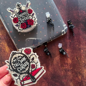 Op de afbeelding: Een doorzichtige telefoonhoes met twee decoratieve pop-up grips en zwarte rozen oorbellen. Eén grip heeft de tekst "Enemies to Lovers" met rozen en zwaarden. De andere heeft "My Type is Tall Dark and Cursed".