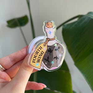 May include: A clear, potion-shaped acrylic charm with a cork stopper and a brown liquid inside. The charm has a tag that reads "FOURTH WING" and features illustrations of bats and stars. The charm is held up by a clear loop.