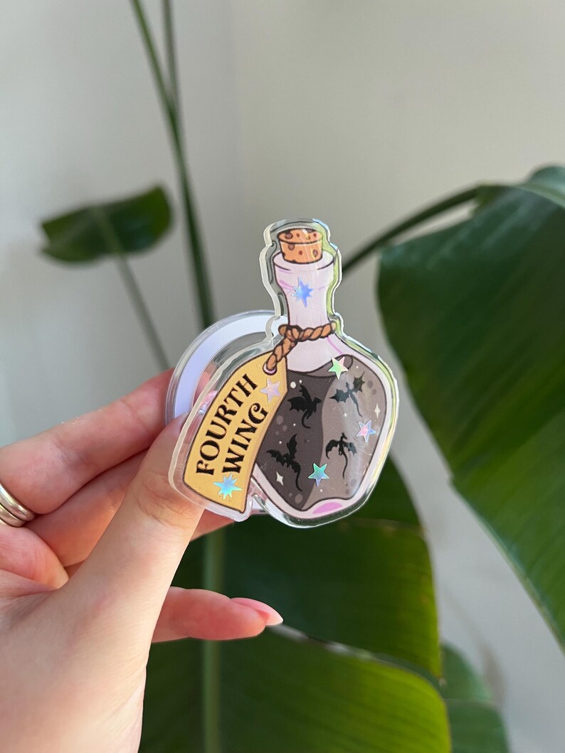 May include: A clear, potion-shaped acrylic charm with a cork stopper and a brown liquid inside. The charm has a tag that reads "FOURTH WING" and features illustrations of bats and stars. The charm is held up by a clear loop.
