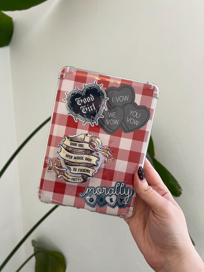 May include: A clear tablet case with a red and white gingham pattern. The case is decorated with several stickers, including a black heart with "Good Girl" and three gray heart-shaped stickers that say "I Vow, We Vow, You Vow". Other stickers include "Open Wider, Baby" and "Morally Grey".