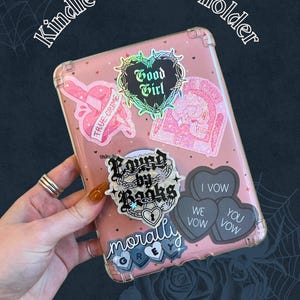 May include: A pink Kindle and phone holder with various stickers. The stickers include phrases like "True Crime," "Good Girl," and "Found by Books." The holder is decorated with heart and skull designs, and the words "I Vow, We Vow, You Vow" are also visible.