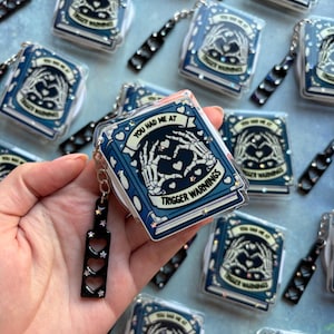 May include: A rectangular blue keychain featuring a graphic of skeleton hands forming a heart shape. The text reads YOU HAD ME AT TRIGGER WARNINGS. It includes a black charm with three heart-shaped cutouts and small star-shaped glitter accents.