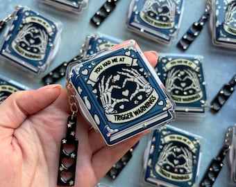 May include: A rectangular blue keychain featuring a graphic of skeleton hands forming a heart shape. The text reads YOU HAD ME AT TRIGGER WARNINGS. It includes a black charm with three heart-shaped cutouts and small star-shaped glitter accents.
