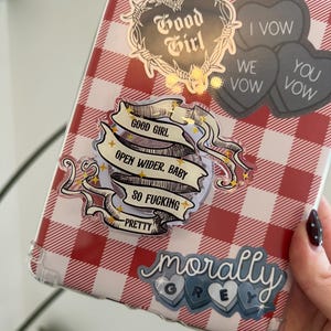 May include: A clear phone case with a red and white checkered pattern. The case is decorated with stickers that say "Good Girl", "Open Wider, Baby, So Fucking Pretty", "I Vow, We Vow, You Vow", and "Morally Grey".