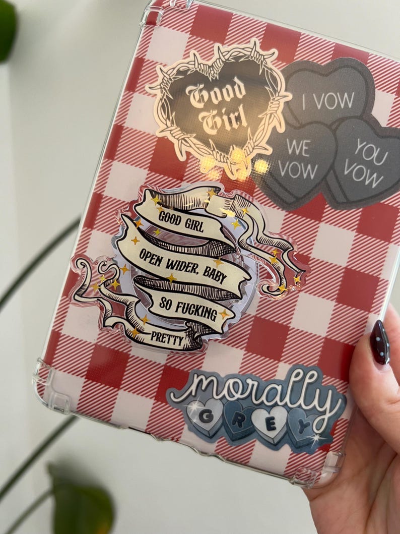 May include: A clear phone case with a red and white checkered pattern. The case is decorated with stickers that say "Good Girl", "Open Wider, Baby, So Fucking Pretty", "I Vow, We Vow, You Vow", and "Morally Grey".