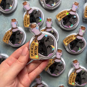 Puede incluir: Una colección de pines de acrílico con forma de frasco de poción que contiene un líquido oscuro con siluetas de dragones voladores y una luna creciente. Cada frasco tiene una etiqueta beige con el texto Fourth Wing y estrellas holográficas.