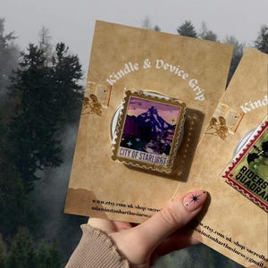 Puede incluir: Un agarre para Kindle y dispositivo con un dise&ntilde;o de paisaje monta&ntilde;oso y el texto "CITY OF STARLIGHT". El agarre est&aacute; adherido a una tarjeta marr&oacute;n con el texto "Kindle & Device Grip". El fondo muestra un bosque.