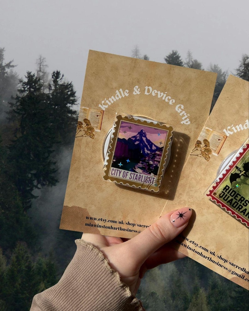 Puede incluir: Un agarre para Kindle y dispositivo con un dise&ntilde;o de paisaje monta&ntilde;oso y el texto "CITY OF STARLIGHT". El agarre est&aacute; adherido a una tarjeta marr&oacute;n con el texto "Kindle & Device Grip". El fondo muestra un bosque.