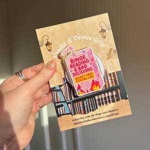 May include: A clear device grip with the text "Binge Reading & Bad Decisions" in pink and red, shaped like a juice box. The grip is attached to a brown card with a bookshelf illustration and the text "Kindle & Device Grip".