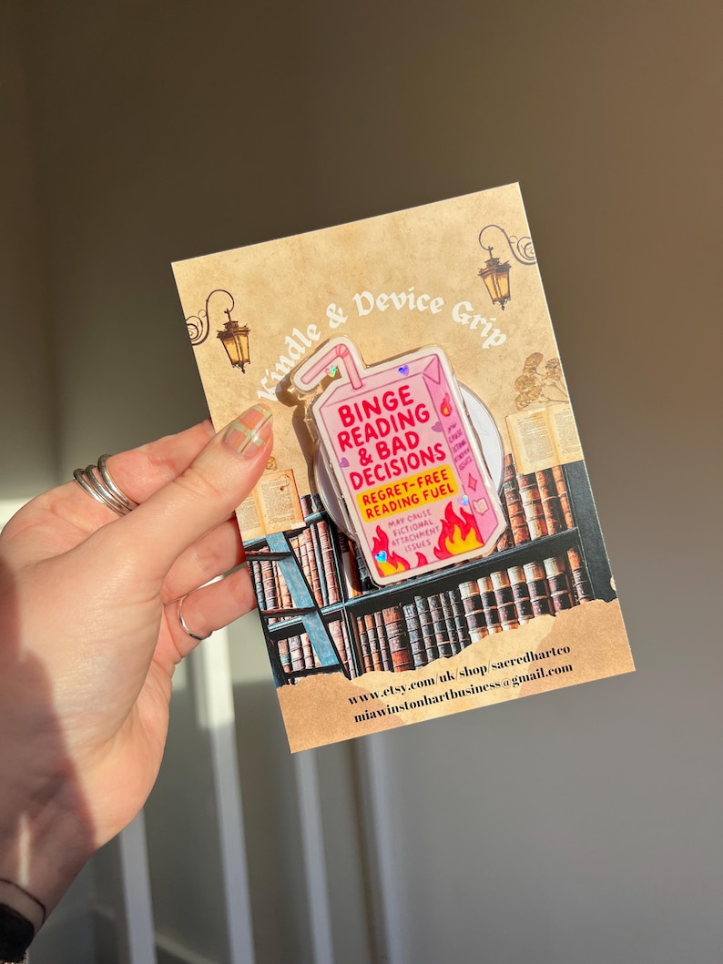 May include: A clear device grip with the text "Binge Reading & Bad Decisions" in pink and red, shaped like a juice box. The grip is attached to a brown card with a bookshelf illustration and the text "Kindle & Device Grip".