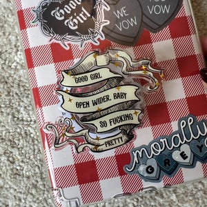 May include: A clear phone case with a red and white checkered pattern, adorned with various stickers. Stickers include a heart-shaped design with the words "Good Girl" and a banner that reads "Open Wider, Baby, So Fucking Pretty."