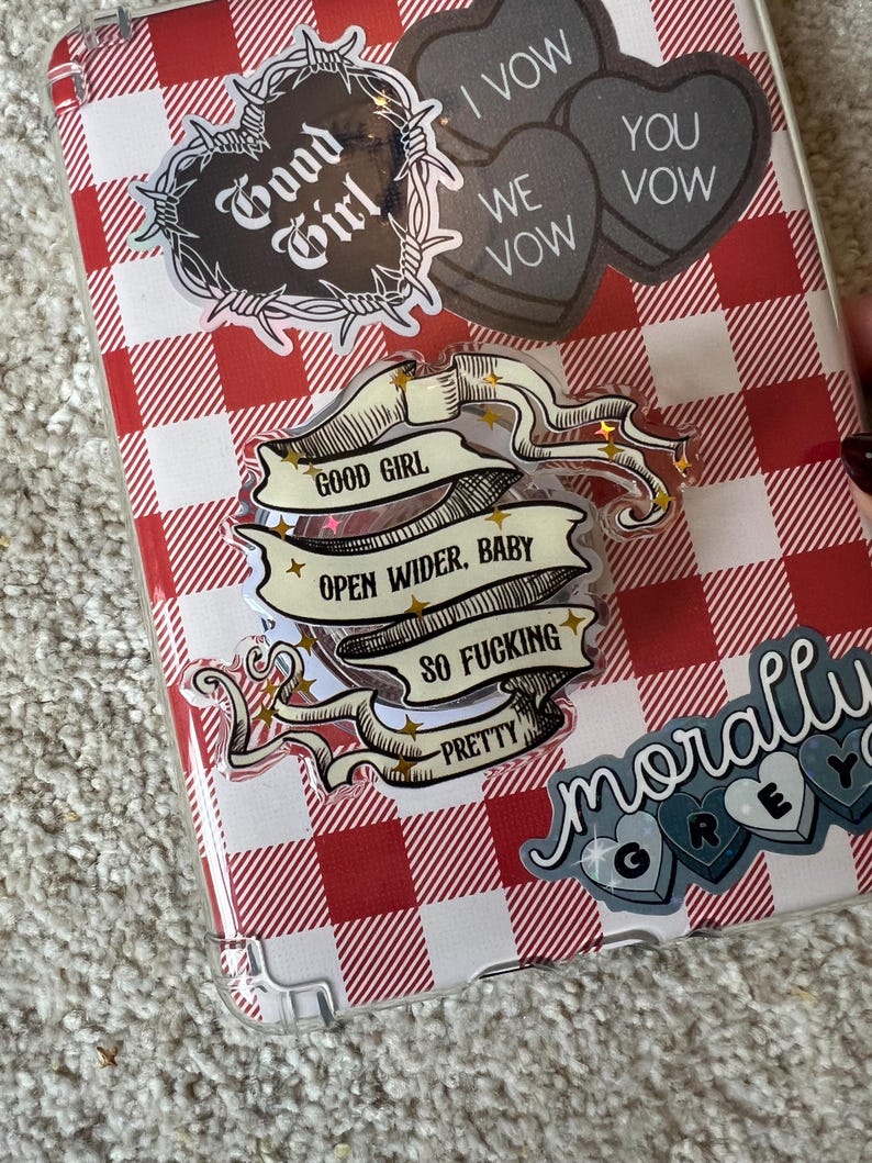 May include: A clear phone case with a red and white checkered pattern, adorned with various stickers. Stickers include a heart-shaped design with the words "Good Girl" and a banner that reads "Open Wider, Baby, So Fucking Pretty."