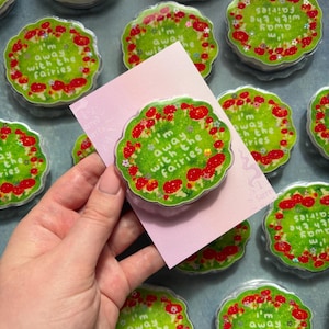 May include: A circular acrylic pin featuring a green background with a border of red mushrooms and small white flowers. The text "I'm away with the fairies" is printed in white in the centre. The pin is attached to a light pink backing card.