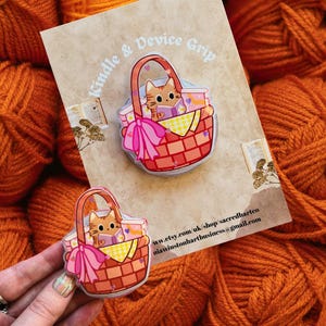 May include: A clear device grip with a cartoon cat in a picnic basket design. The basket is red with a pink bow and yellow checkered blanket. The text "Kindle & Device Grip" is visible.