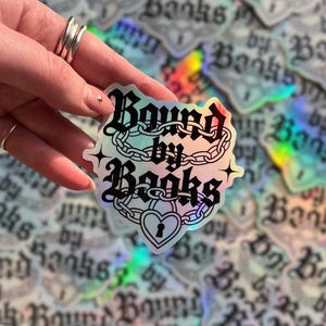 May include: Holographic sticker with the text "Bound by Books" in a gothic font, featuring chain and heart lock illustrations. The sticker has a shimmering, iridescent finish and is held by a hand. The background is filled with similar stickers.