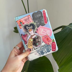 May include: A clear phone case decorated with various stickers. The stickers include a potion bottle, hearts with text, a book, and the words "TOUCH HER AND DIE book club". The case is held by a hand.