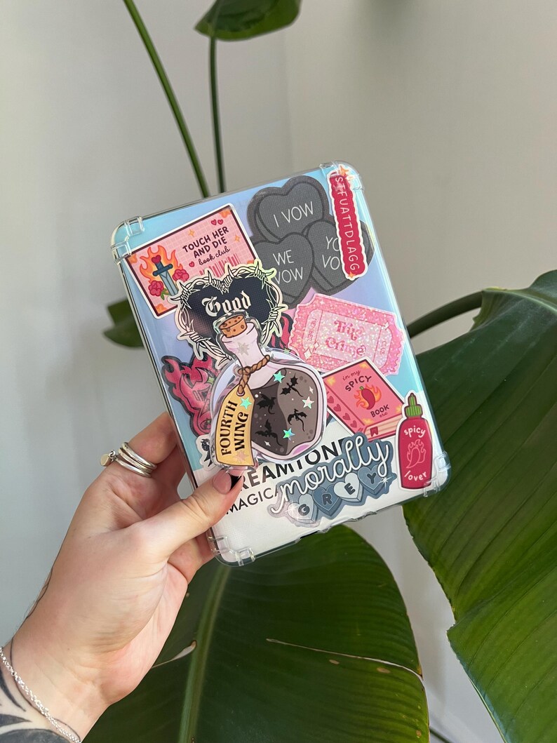 May include: A clear phone case decorated with various stickers. The stickers include a potion bottle, hearts with text, a book, and the words "TOUCH HER AND DIE book club". The case is held by a hand.
