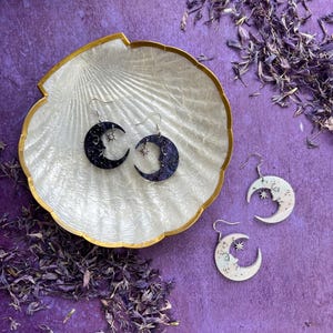 May include: A pair of crescent moon earrings with stars, one pair is black with silver stars and the other is white with gold stars. The earrings are displayed on a white seashell dish.