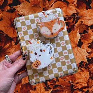 Può includere: Due spille in acrilico raffiguranti una volpe e un fantasma, ciascuna disegnata come una tazza di caffè con panna montata e decorazioni a tema autunnale. Le spille poggiano su una custodia a scacchi oro e bianca, su uno sfondo di foglie autunnali arancioni.