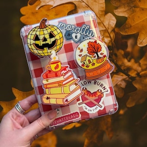 May include: A clear phone case with a red and white gingham pattern, decorated with autumn-themed stickers. Stickers feature a carved pumpkin, a snow globe with a house and tree, a stack of books with a teacup, and a heart with the text "I'm on fire."