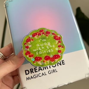 May include: A clear phone case with a light blue and pink gradient. A green, circular sticker with red mushroom illustrations and the text "I'm away with the fairies" is attached. The text "DREAMTONE MAGICAL GIRL" is printed on the case.