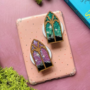 May include: Two wooden, arched window-shaped pins. One pin features a green and white starry night scene over a mountain range. The other pin has a purple and pink starry night scene over a mountain range. Both pins are on white circular bases.