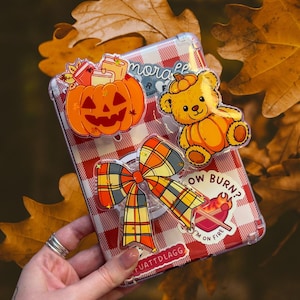 May include: A clear phone case with a red and white checkered pattern, adorned with fall-themed stickers. Stickers include an orange pumpkin, a teddy bear, and a plaid bow. The case also has the text "I'M ON FIRE".
