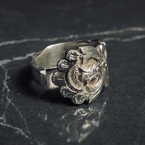 Puede incluir: Un anillo ajustable plateado con un diseño ornamentado. El anillo presenta un medallón central con un relieve detallado, rodeado de motivos florales y de hojas. La banda del anillo es abierta y ajustable.