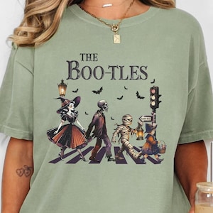以下が含まれることがあります： 横断歩道を歩く魔女、ゾンビ、ミイラ、そして幽霊に扮した子供のグラフィックが描かれた緑色のTシャツ。グラフィックの上に「THE BOO-TLES」という文字が書かれている。