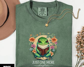 Może przedstawiać: Zielona koszulka w kolorze mchu z ilustracją żaby czytającej książkę, otoczonej kwiatami. Tekst "JUST ONE MORE chapter" znajduje się pod żabą. Złoty naszyjnik z zawieszką w kształcie gwiazdy jest na koszulce.
