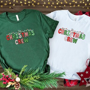 May include: Two green and white t-shirts with the text "Christmas Crew" in red, green, and white letters with a string of Christmas lights above the text.