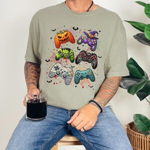 Op de afbeelding: Een kaki T-shirt met een Halloween-thema met meerdere videogamecontrollers met griezelige details zoals pompoenen, vleermuizen en spinnen.