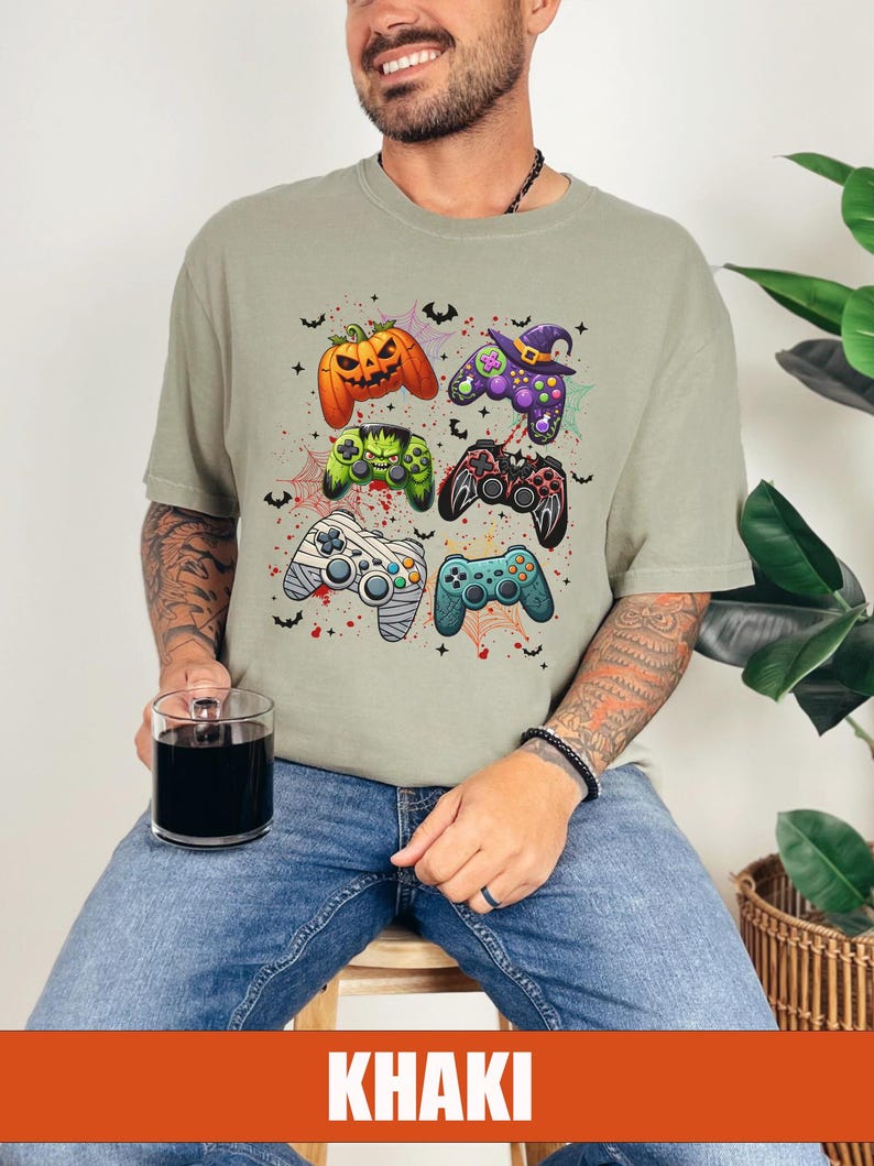Op de afbeelding: Een kaki T-shirt met een Halloween-thema met meerdere videogamecontrollers met griezelige details zoals pompoenen, vleermuizen en spinnen.