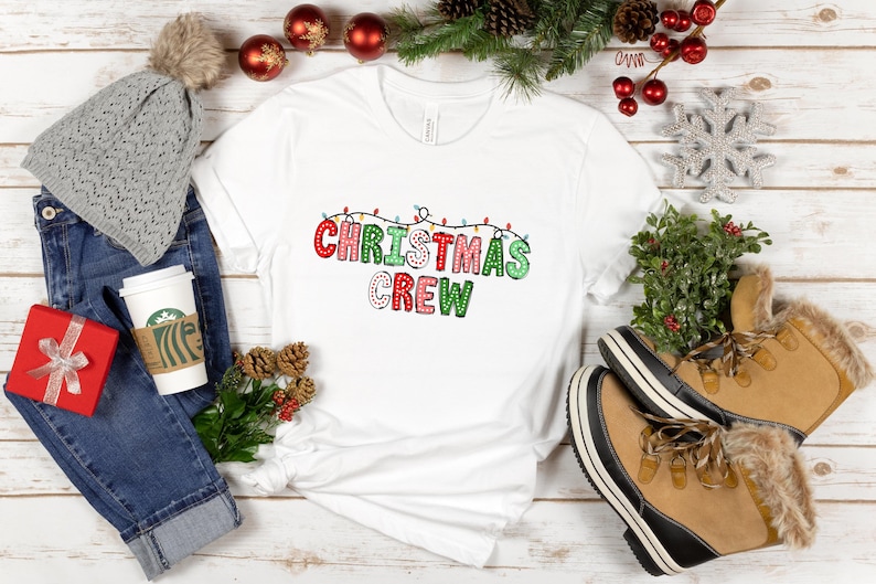 May include: White t-shirt with the text "Christmas Crew" in red, green, and white letters with white polka dots. The letters are outlined in black and have small white lights strung above them.