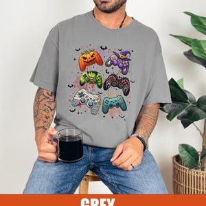 Op de afbeelding: Een grijs T-shirt met een Halloween-thema met verschillende videogamecontrollers versierd met griezelige elementen zoals pompoenen, vleermuizen en een heksenhoed.