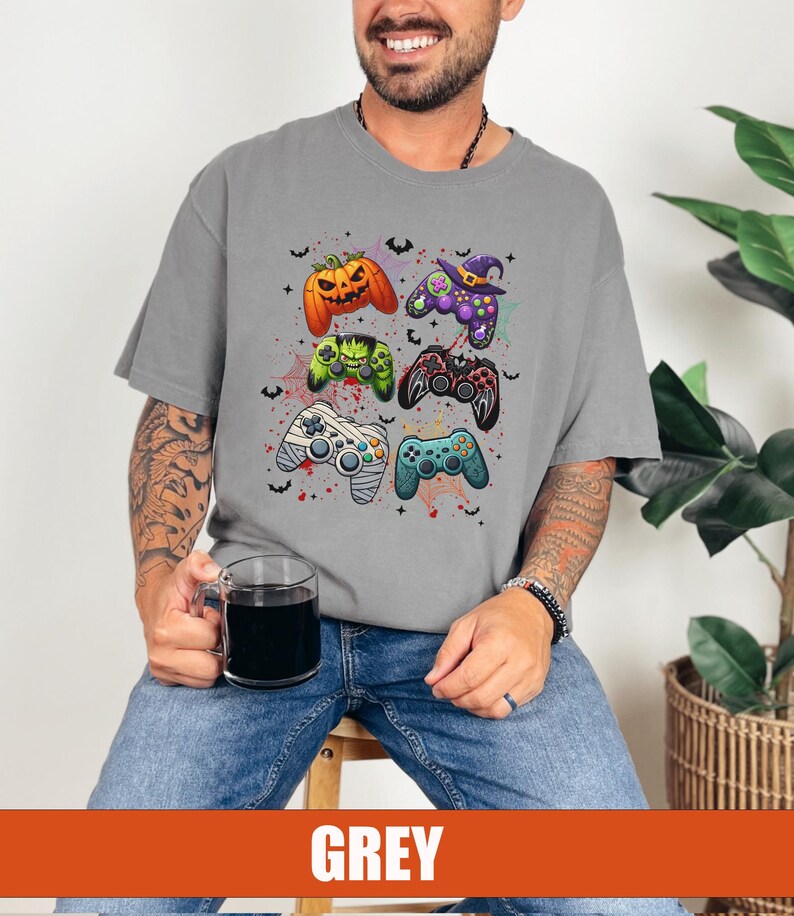 Op de afbeelding: Een grijs T-shirt met een Halloween-thema met verschillende videogamecontrollers versierd met griezelige elementen zoals pompoenen, vleermuizen en een heksenhoed.