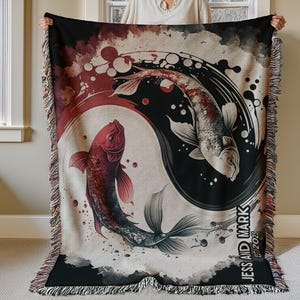 Op de afbeelding: Een geweven tapijtdeken met een yin yang-ontwerp met twee koi-vissen. De vissen zijn rood en wit met zwarte accenten. De achtergrond is een aquarelverf in rood, zwart en wit. De tekst "Jess and Mark Est. 2023" is afgedrukt in de rechterbenedenhoek.