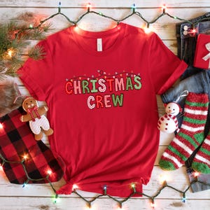May include: Red t-shirt with the text "Christmas Crew" in colorful letters with string lights above the text.