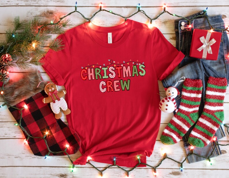 May include: Red t-shirt with the text "Christmas Crew" in colorful letters with string lights above the text.
