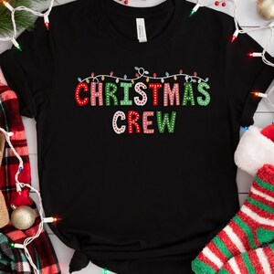 May include: Black t-shirt with the text "Christmas Crew" in colorful letters with white string lights above the text. The letters are decorated with small white dots.