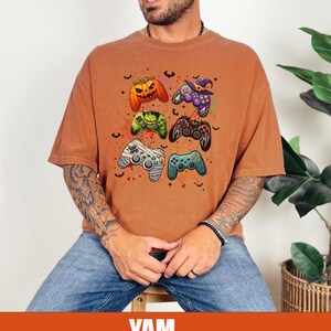 Op de afbeelding: Een oranje T-shirt met een afbeelding van Halloween-videogamecontrollers. De controllers zijn versierd met pompoenen, vleermuizen en heksenhoeden.
