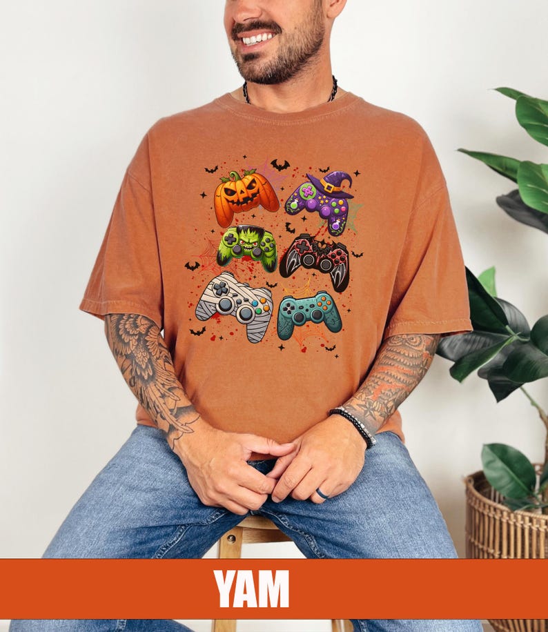 Op de afbeelding: Een oranje T-shirt met een afbeelding van Halloween-videogamecontrollers. De controllers zijn versierd met pompoenen, vleermuizen en heksenhoeden.