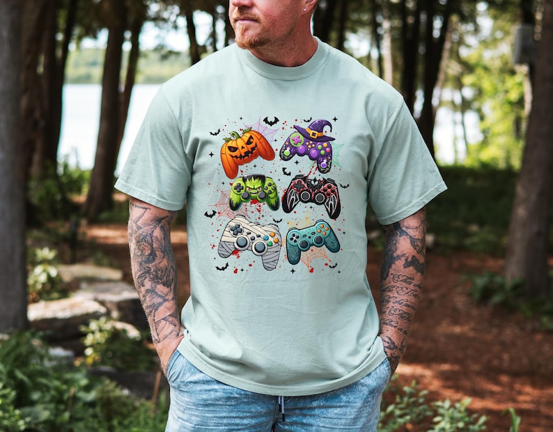 Op de afbeelding: Een lichtblauw T-shirt met een afbeelding van zes verschillende Halloween-videogamecontrollers. De controllers zijn versierd met pompoenen, geesten, vleermuizen en een heksenhoed.
