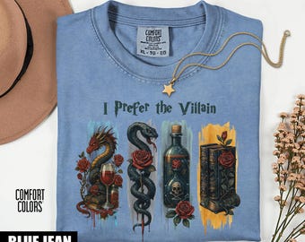 Op de afbeelding: Een lichtblauw Comfort Colors T-shirt met de tekst "I Prefer the Villain". Het shirt heeft illustraties van een draak, slang, drankfles en boeken, allemaal versierd met rozen. Een gouden ketting met een sterhanger is over het shirt gedrapeerd.