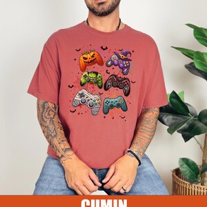 Op de afbeelding: Een rood T-shirt met een afbeelding van vijf verschillende gekleurde videogamecontrollers. De controllers zijn versierd met Halloween-thema's, waaronder een pompoen, een heksenhoed en een mummie.