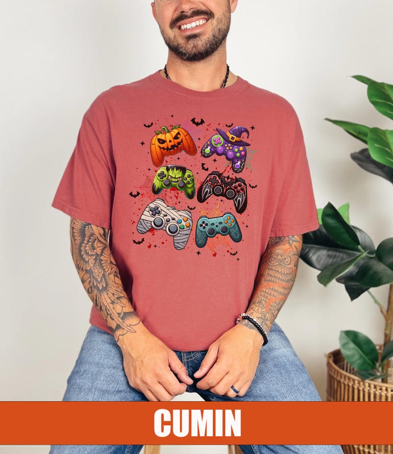 Op de afbeelding: Een rood T-shirt met een afbeelding van vijf verschillende gekleurde videogamecontrollers. De controllers zijn versierd met Halloween-thema's, waaronder een pompoen, een heksenhoed en een mummie.