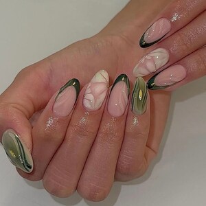 May include: Close-up of a hand with almond-shaped nails painted with a light pink base and green and gold abstract designs.